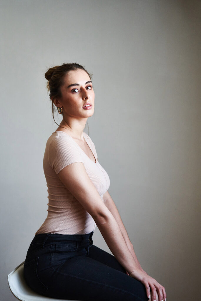 Marintia Bustos a model with a radiant smile sits gracefully on a white stool against a neutral backdrop. She wears a soft pink top and dark jeans and her earrings shimmer with elegance.
